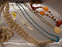 Five necklaces and a matching beige bracelet laid out on white fabric, featuring various bead styles and pendants.