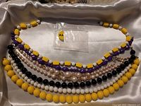 Photo showing six beaded necklaces in various colors including yellow, purple, black, white, and pearl-like beads arranged on fabric background.