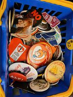 Top view of mixed vintage buttons inside blue plastic basket showing various slogans and cartoon graphics from the 80s and 90s.