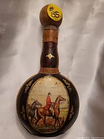 Front view showing leather-wrapped decanter with painted horseman and horses in countryside scene, gold embossed designs