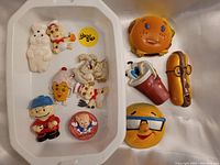 Photo of 11 novelty 1980s/90s fridge magnets arranged in a white tray and on white surface, colorful character and food item designs, plastic and rubber material