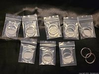 Eight pairs of hoop earrings individually packaged in clear plastic bags, arranged on a black surface with one pair out of the bag for display.