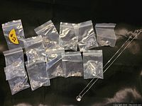 Eleven separate sealed plastic bags each containing components of the necklaces scattered on a dark fabric background, showing multiple silver chains and pendants.