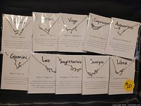 Photo showing 10 individually packaged silver tone zodiac necklaces, each with a pendant and informational card describing the zodiac sign and date range.