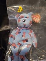 Blue plush bear with American flag pattern, sealed in a clear plastic protective bag, showing the TY Beanie Baby heart tag attached to the ear and a small yellow sticker labeled #30.