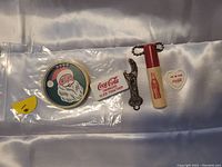 All five vintage Coca-Cola items arranged on a cloth background showing front faces including Santa dexterity game, noise clicker, bottle opener, license key chain, and bookmark