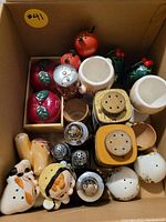 Top view of assorted salt and pepper shakers including fruit and character designs, and wooden and ceramic egg cups.