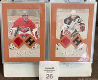 Photo of two 2004 game-used jersey cards featuring Jose Theodore and Chris Drury in Beehive design with patches in brown matting.