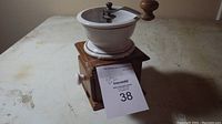 Manual coffee grinder front angle showing porcelain grinding bowl, wooden base drawer with white knob, metal handle with wooden knob.