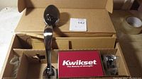 Open box photo showing the metallic handle, Kwikset branded installation instructions/manual, and locked hardware accessories inside the brown shipping box.