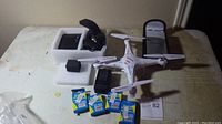 Photo showing the Syma Model X5C drone with four propellers, four lithium-ion batteries in blue and yellow packaging, battery charger, wrist strap holder, and protective pouch on a table.