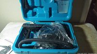 Open blue plastic case showing black Turbo Steamer 220 unit in plastic bag, flexible hose, and various attachments packed inside case.