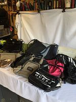 Wide view of 9 purses and bags on white surface with some overlapping and leaning together. Colors vary from black to red to white and beige.
