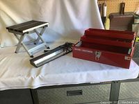 3 items displayed on a white cloth: red Sears Craftsman toolbox, light bar with power cord, and folded aluminum seat stool.
