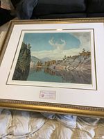 Full view of framed lithograph showing river landscape with rocky shores and trees reflecting in water under a blue sky.