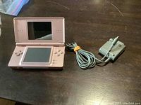 Pink Nintendo DS Lite handheld game system open showing screens and control buttons, with grey power adapter cable placed next to it.