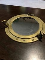 Round brass ship's porthole with mirror replacing the glass, shown from an overhead angle on a dark wood surface.