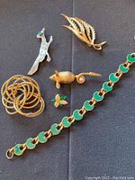 Photo showing mixed costume jewelry including leaf-shaped brooch, mouse brooch, bird brooch, floral pin, linked bracelet, and set of expansion rings on black background.