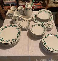 Full set of Christmas dinnerware laid out on table including plates, bowls, cups, saucers, teapot, gravy boat, sugar dish, platter, and serving bowl all with holly and berry rim pattern.