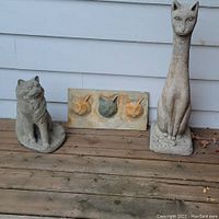 Three concrete cat statues on wood porch floor against white siding: tall slim cat statue, sitting cat statue, and rectangular cat head wall hanging.