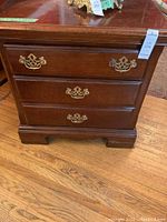 Front view of one wooden end table showing three drawers with brass handles and stepped lower base.