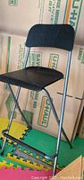 Side view of one IKEA folding bar chair with black plastic seat and backrest and silver metal frame, positioned on interlocking rubber floor mats and in front of moving boxes.