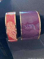 Photo of two enamel cuff bracelets, red with gold and purple with lighter pattern, displayed on black bracelet stand.