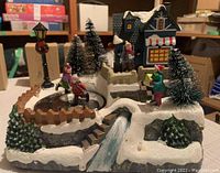 Winter village scene with snow-covered house, pine trees, stone bridge, fence, and ice skater figurines on frozen pond.