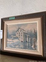 Framed oil painting showing snowy mill scene by water with reflection, housed in brown wooden frame with beige mat.