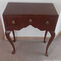 Front-facing photo of the dark brown wooden side table with three drawers, brass ring handles, and decorative carved apron on four cabriole legs.