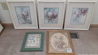 Five framed art pieces arranged on floor including four white floral/botanical prints and one green framed decorative print.