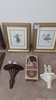 Six framed food and fruit art prints including two gold frames and four dark brown wood frames with burgundy mats. Also shown are Precious Moments nativity collection figurines in original box, plus loose figurines including camel and bride and groom, and two decorative wall shelves (one dark wood and one white cherub design).