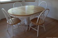Photo of the wooden oval kitchen table with four chairs positioned around it inside a room with wooden flooring and windows with blinds.