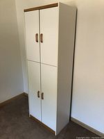 Closed view of white tall storage cabinet with four doors and wooden handles, wood trim on top and bottom.