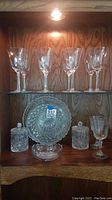 Wooden cabinet shelf holding 5 wine glasses, round crystal plate, sugar bowl and creamer, small crystal bowl and wine glass on lower shelf under light