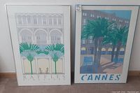 Photo of two large framed travel prints Raffles Singapore and Cannes on wall, showing architectural and palm tree scenes in muted colors with titles at bottom