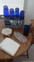 Photo showing blue kitchen canisters, boxed crystal wine glasses, blue pitcher, oval crystal serving dish, cream tablecloth, and divided serving dish on wooden table