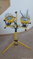 Full view of the yellow Workforce double halogen stand light with two light heads and tripod base, including power cord and extra bulbs holder.