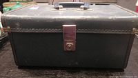 Front view of the black hard-sided rectangular case with metal latch and handle on the top.