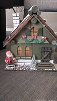 Front view of the vintage wooden Christmas decoration house with light-up windows, Santa figure, and snow-covered base.