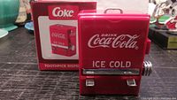 Front view of red Coca-Cola toothpick dispenser with white logo, dispensing levers, and chrome knob, with original box behind