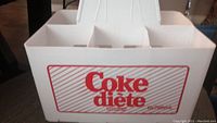 Front view of white plastic bottle carrier with red 'Coke diete' logo and diagonal stripes