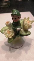 Front angle showing the pixie sitting among yellow flowers and green leaves on a white base. The pixie wears a green outfit and hat.