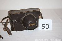 Closed view of vintage Argus camera inside original brown leather case with strap. The camera lens cover prominently marked "argus".