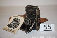 Front angled view of the Balda Baldina camera with lens extended and instruction manual beside it, showing lens and shutter details.