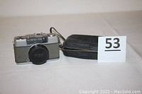 Front view of Olympus PEN camera with lens cap attached and case beside it.
