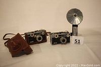 Two Argus C3 cameras, brown leather case, and silver flash attachment displayed on neutral background.