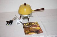 Wide view showing mustard yellow fondue pot on metal and wooden stand, assorted fondue forks with colored tips, and 1970 Better Homes and Gardens fondue cookbook.
