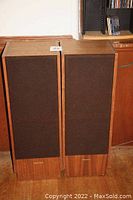 Pair of Realistic Optimus T-100 tower speakers with brown fabric grilles on, standing side by side on wood floor.