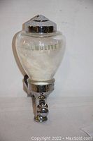 Front view of soap dispenser showing Palmolive logo on glass reservoir and chrome top with valve at bottom.